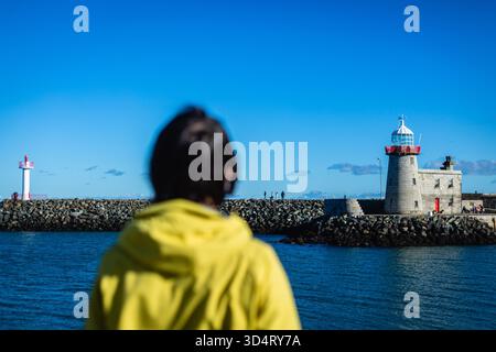 vista posteriore di una persona in giacca gialla che guarda la vista panoramica sul mare e sul faro Foto Stock