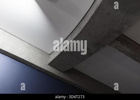 Un sorprendente dettaglio interno caratterizzato da travi curve in cemento che attraversano pannelli bianchi e una parete blu, che mette in risalto l'architettura moderna, le trame e un i Foto Stock