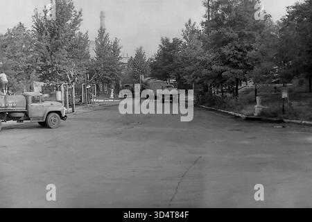 Una foto d'archivio in bianco e nero degli anni '1970 mostra una vista generale del deposito petrolifero di Sloviansk, la RSS Ucraina. Diversi autocisterne sovietici sono parcheggiati lungo una strada asfaltata costeggiata da alti alberi frondosi, con un lavoratore visibile su una cisterna. Sullo sfondo, un'alta cisterna industriale sorge sopra la struttura, definendo il paesaggio. Questa immagine cattura la logistica di routine e la miscela unica di industria e natura all'interno del pacifico Donbas durante l'era sovietica. Foto Stock