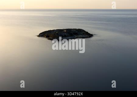 Uno scatto focalizzato e simmetrico di una formazione rocciosa scura isolata che riposa nell'acqua tranquilla e cristallina. La consistenza liscia della superficie del mare contrasta Foto Stock