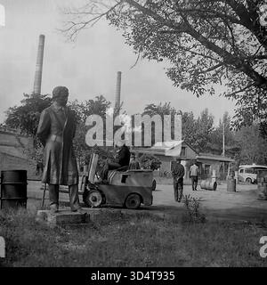 Una foto d'archivio in bianco e nero degli anni '1970 mostra il paesaggio industriale del deposito petrolifero di Sloviansk, la RSS Ucraina. In primo piano, una statua di epoca sovietica si erge sull'erba. A metà strada, un lavoratore gestisce un piccolo carrello elevatore a tre ruote, mentre altri lavoratori si dirigono verso un edificio di pubblica utilità. Diverse botti di metallo sono a terra, e un furgone bianco è parcheggiato nelle vicinanze. Sullo sfondo, due alte cisterne industriali si innalzano sopra gli alberi, definendo il paesaggio. Questa scena cattura il lavoro quotidiano e l'atmosfera industriale del tranquillo Donbas. Foto Stock