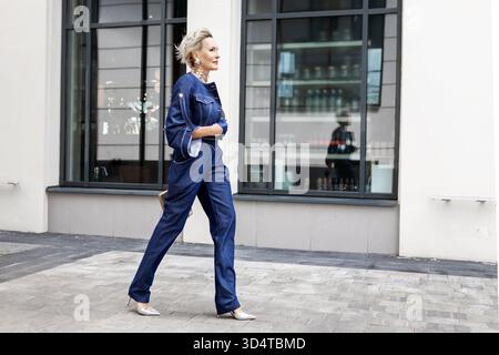 Donna elegante e sicura di sé che cammina all'aperto sul marciapiede della città indossando una giacca in denim alla moda e jeans con tacco alto. Elegante modella da donna d'affari, mod Foto Stock