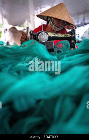Veduta aerea di una sarta in un cappello conico che lavora meticolosamente alla sua macchina da cucire, circondata da un mare di vivace tessuto verde, il Vietnam. Foto Stock