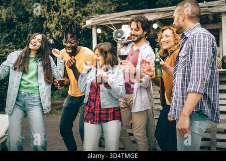 Un gruppo felice e multietnico di sei giovani amici balla, beve e ascolta la musica da un boombox a una festa all'aperto nel cortile. Foto Stock