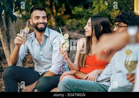 Un gruppo felice e multietnico di giovani amici siede all'aperto, parlando e bevendo cocktail a una festa in giardino estiva. Foto Stock