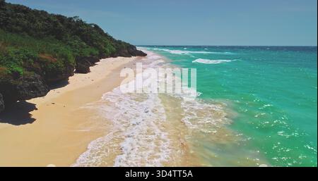 Vista aerea mozzafiato che cattura le onde turchesi dell'oceano che si infrangono dolcemente su una spiaggia di sabbia incontaminata, circondata da una lussureggiante vegetazione verde sull'isola di Sumba, Indonesia Foto Stock