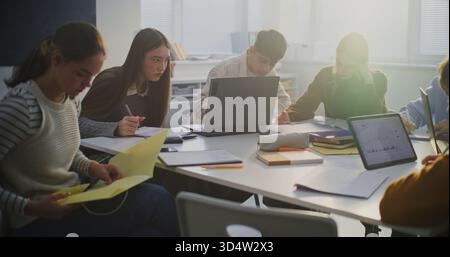 Gruppo di studenti riuniti nell'area di lavoro condivisa, collaborando attivamente all'assegnazione utilizzando notebook, tablet e note cartacee. Concetto di risoluzione dei problemi cooperativi e Modern Classroom Teamwork. Foto Stock