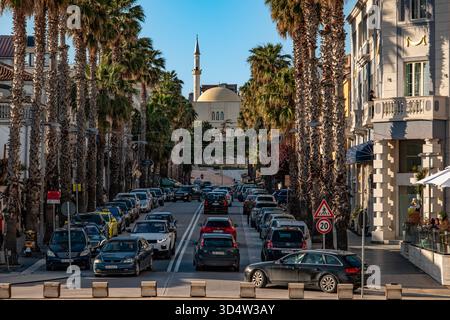 Passeggiata con palme e molti negozi e ristoranti nel centro di Durazzo, Albania. C'è traffico intenso sulla strada e le auto sono parcheggiate lungo la strada. Foto Stock