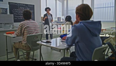 Insegnante femminile si trova vicino alla lavagna che spiega l'assegnazione della codifica al gruppo di studenti in Bright Classroom. Concetto di istruzione moderna, apprendimento interattivo, lavoro di squadra creativo in ambiente scolastico. Foto Stock
