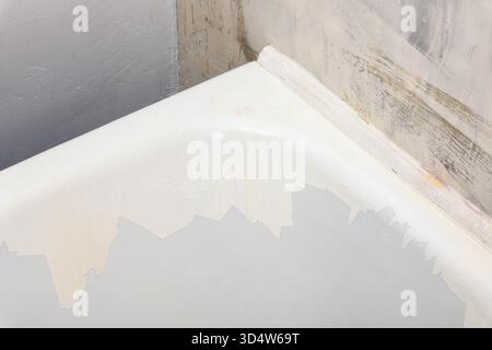 Primo piano di una vasca da bagno con vernice bianca sbucciata contro pareti non rifinite e ruvide. Ideale per progetti di ristrutturazione, riparazione o miglioramento della casa. Foto Stock