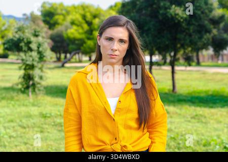 Donna seria in camicia gialla che guarda la macchina fotografica all'aperto con un'espressione neutra Foto Stock