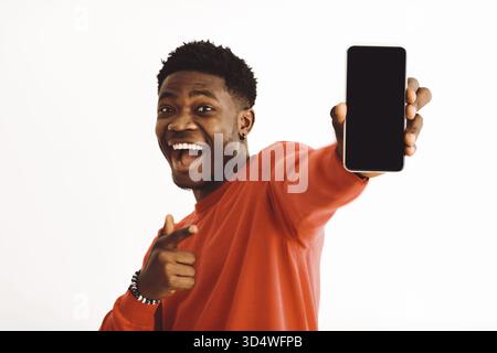 Young Man sorride e presenta lo smartphone in un allegro ambiente interno Foto Stock