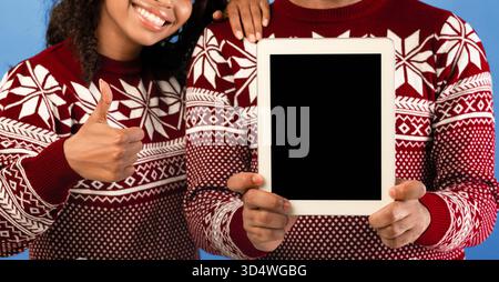Concetto di vendite di Natale. Uomo nero che tiene il tablet con schermo vuoto e donna che mostra il pollice su sfondo blu Foto Stock