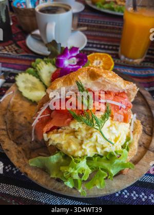 Croissant per la colazione gourmet, ricco di salmone affumicato, cremose uova strapazzate, lattuga croccante e aneto. Servito su un piatto rustico di legno con coff caldo Foto Stock