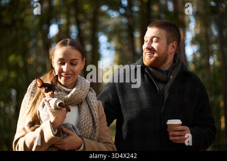 Giovane donna caucasica che tiene in mano un piccolo cane, mentre si trova accanto al giovane uomo caucasico, sorride e trasporta tazza di caffè usa e getta, all'aperto nella foresta durante il giorno Foto Stock