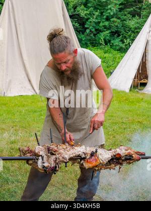 Bøstad, Norvegia - 3 agosto 2023: L'uomo in costumi popolari medievali arrostisce un maiale allo spiedo durante il Festival vichingo presso il Museo vivente dei Vichinghi in Foto Stock