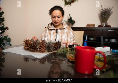 Una donna lavora su decorazioni pinecone in un accogliente tavolo di casa, creando decorazioni natalizie con accenti natalizi e accenti natalizi sullo sfondo. Foto Stock