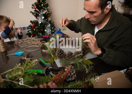Un uomo e un bambino realizzano una corona natalizia con pinne e rametti sempreverdi a un tavolo da pranzo, mentre un albero di Natale brilla sullo sfondo. Foto Stock