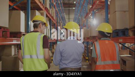 Walking 3 warehouse workers wearing yellow hard hats and vests inspecting in warehouse with tablet Foto Stock