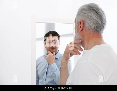 Uomo anziano che si vede giovane allo specchio. Concetto di personalità, passato e futuro, flusso del tempo, età Foto Stock