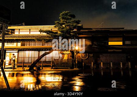 Vista della strada bagnata che riflette la luce dorata degli edifici tradizionali sotto un cielo tempestoso, con un albero di pino davanti, Takayama, Gifu, Giappone. Foto Stock