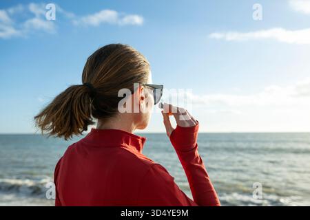 Foto del profilo di una donna sportiva con un abbigliamento sportivo rosso, occhiali da sole e auricolari wireless che guardano l'oceano Foto Stock