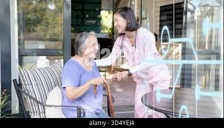 Donna asiatica sorridente che tiene la canna sul patio, caregiver con stetoscopio appoggiato Foto Stock
