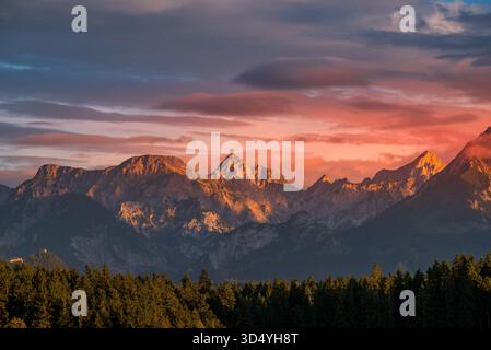 Vista delle maestose vette delle montagne baciate da un vibrante tramonto, che proietta una calda luce sulla lussureggiante foresta verde sottostante, Nesselwang, Bayern, Germania. Foto Stock