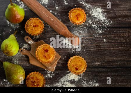 Le crostate di pere appena sfornate siedono su un tagliere di legno, circondate da pere e farina. Un mattarello si trova nelle vicinanze, creando un'atmosfera calda e invitante Foto Stock