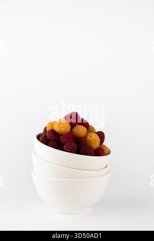 Le bacche sono disposte splendidamente in tre ciotole bianche sovrapposte, con lamponi rossi vibranti e frutti dorati luminosi contro un ba pulito e minimale Foto Stock