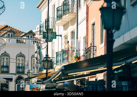 Lagos, Portogallo, 8 novembre 2025. Una donna si erge su un balcone al secondo piano che si affaccia su una strada vivace fiancheggiata da edifici color pastello. Foto Stock