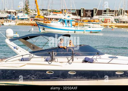 Lagos, Portogallo, 8 novembre 2025. Un uomo e una donna si rilassano su uno yacht moderno vicino al porto turistico, con barche e cantieri sullo sfondo. Foto Stock