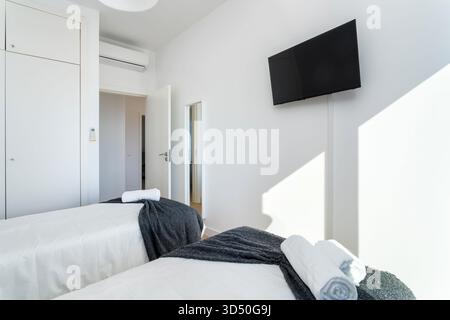 Due letti singoli con morbide lenzuola e asciugamani sono disposti in una camera da letto luminosa e minimalista dotata di un grande televisore a parete. La camera e' accogliente e. Foto Stock