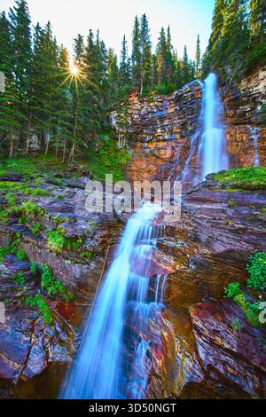 Cascata che scende lungo Rocky Cliff con Sunburst attraverso la foresta di pini Foto Stock