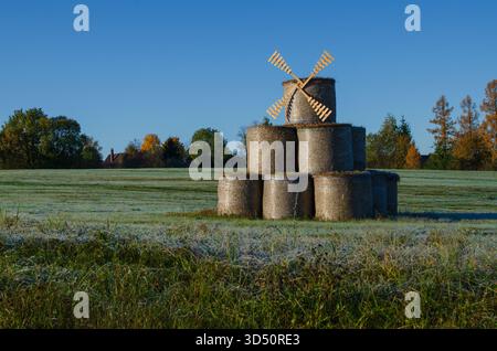 Un mulino per paglia costruito con balle di fieno appena tagliato, l'erba giovane ricoperta di gelo mattutino. Un cielo blu e alberi autunnali. Le pulegge scanalate sono impilate Foto Stock