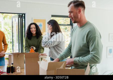 Diversi amici confezionano scatole con dispenser di nastro intorno a un tavolo in legno in una casa luminosa e in uno spazio di copia Foto Stock