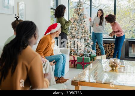 Amici diversi che decorano l'albero di Natale e sistemano scatole regalo avvolte a casa accanto al caminetto Foto Stock