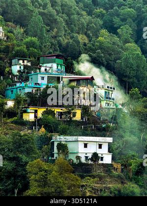 Questa immagine cattura splendidamente un tranquillo villaggio collinare annidato tra fitte foreste verdi. Le case vibranti, dipinte in tonalità turchesi, gialle e bianche, si ergono graziosamente su pendii terrazzati, creando una scena pittoresca tipica delle regioni himalayane o collinari dell'India settentrionale. Le ciuffe di fumo si alzano dolcemente da una delle case, suggerendo fuochi di prima mattina o di sera, un simbolo semplice ma avvolgente della vita rurale quotidiana. La foresta circostante, piena di pini alti e fitte fogliame, avvolge il villaggio in una coperta di tranquillità, facendolo sentire quasi nascosto al Foto Stock