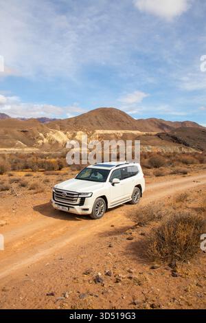 Naryn, Kirghizistan - 13 dicembre 2024: Una Toyota Land Cruiser 300 bianca che guida su una strada sterrata rossa in una valle con colline aride drammaticamente colorate e. Foto Stock