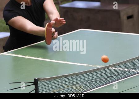 Una donna concentrata che gioca a ping pong all'aperto colpisce una palla arancione su una rete con la pagaia. Questo sport attivo e ricreativo richiede concentrazione e abilità Foto Stock