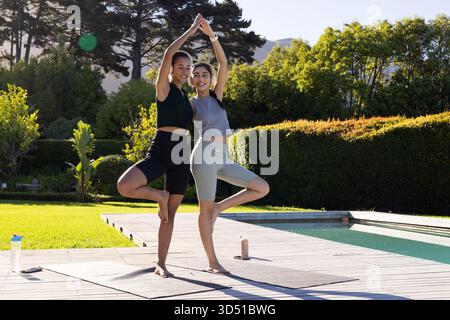 Bilanciare diverse amiche che praticano posa sugli alberi su tappetini da yoga a bordo piscina con bottiglie d'acqua Foto Stock