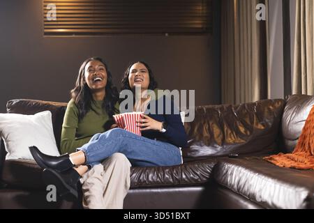 Ridendo diverse amiche che condividono popcorn da un secchio a righe sul divano a casa, con cuscino Foto Stock