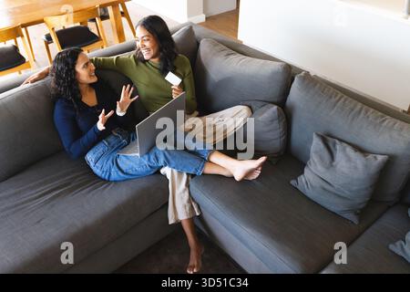 Far sedere diverse amiche che navigano nel negozio online sul computer portatile in salotto, tenendo in mano la carta di credito Foto Stock
