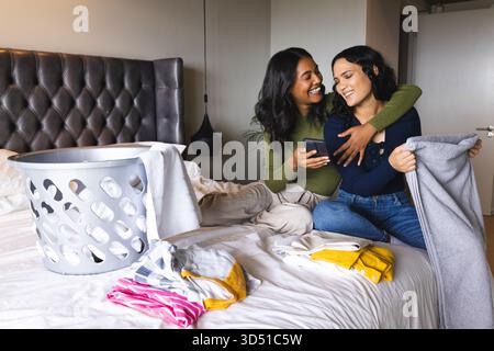 Sorridere due donne sedute insieme sul letto in camera da letto, tenendo in mano smartphone e cestino per la lavanderia grigio Foto Stock
