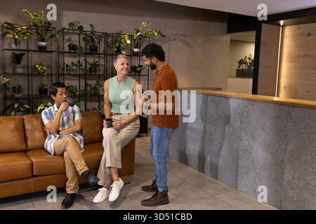 Chiacchierando con colleghi diversi seduti su un divano in pelle nella moderna sala dell'ufficio, con tazze da caffè Foto Stock