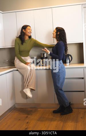 Chiacchierando con diverse amiche che si appoggiano sul piano di lavoro in cucina, con smartphone e macchina per il caffè Foto Stock
