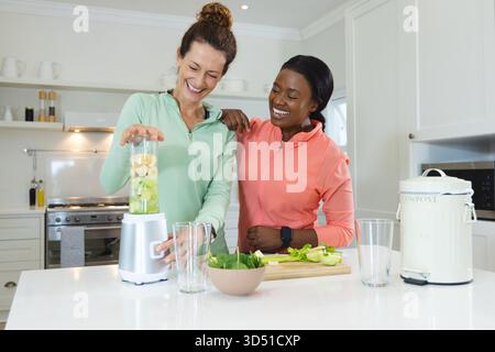 Sorridendo diverse amiche che preparano frullati verdi all'isola della cucina, con frullatore e prodotti Foto Stock