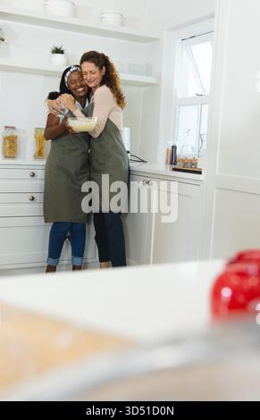 Abbraccia diverse amiche mescolando la pastella nella cucina di casa, con recipiente per mescolare e vasetti di vetro Foto Stock