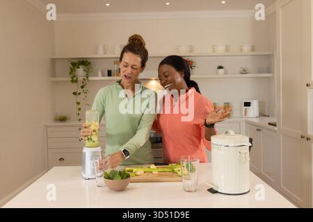 Diverse amiche che mescolano frullati nella cucina della fattoria, con frullatore e spinaci freschi Foto Stock