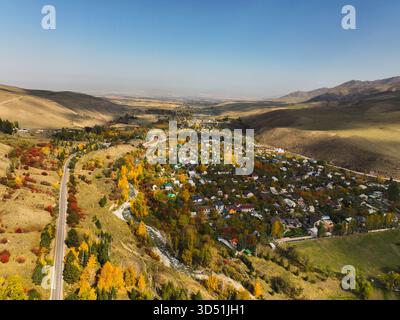 Una vista aerea ad alto angolo di un piccolo villaggio in una valle di montagna, circondato da colline e alberi che mostrano un vivace fogliame autunnale rosso e dorato, con Foto Stock